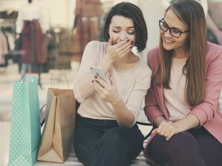 Two shoppers laugh at a phone between their shopping bags