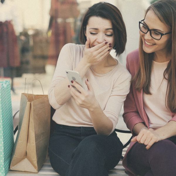 Two shoppers laugh at a phone between their shopping bags